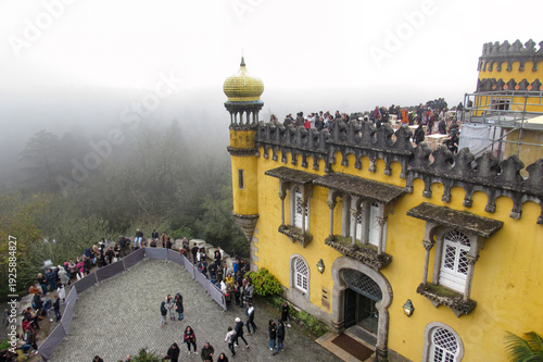 Tourists visiting the colorful terraces of Pena Palace in Sintra, Portugal, with yellow walls, decorative battlements, and panoramic views over the surrounding forest on a misty day.