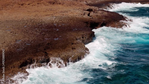 Impresionante vista aérea de las olas rompiendo sobre las rocas rojizas de Aruba, revelando el hermoso paisaje con acantilados y las texturas de las rocas al sol