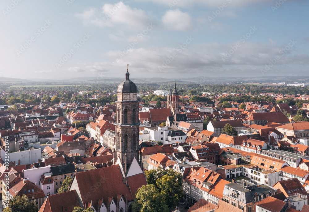 Fototapeta premium Drone cityscape of Goettingen (Göttingen), Germany, with a historic church tower rising above red tiled roofs and the old town stretching toward the horizon under a bright sky with clouds.