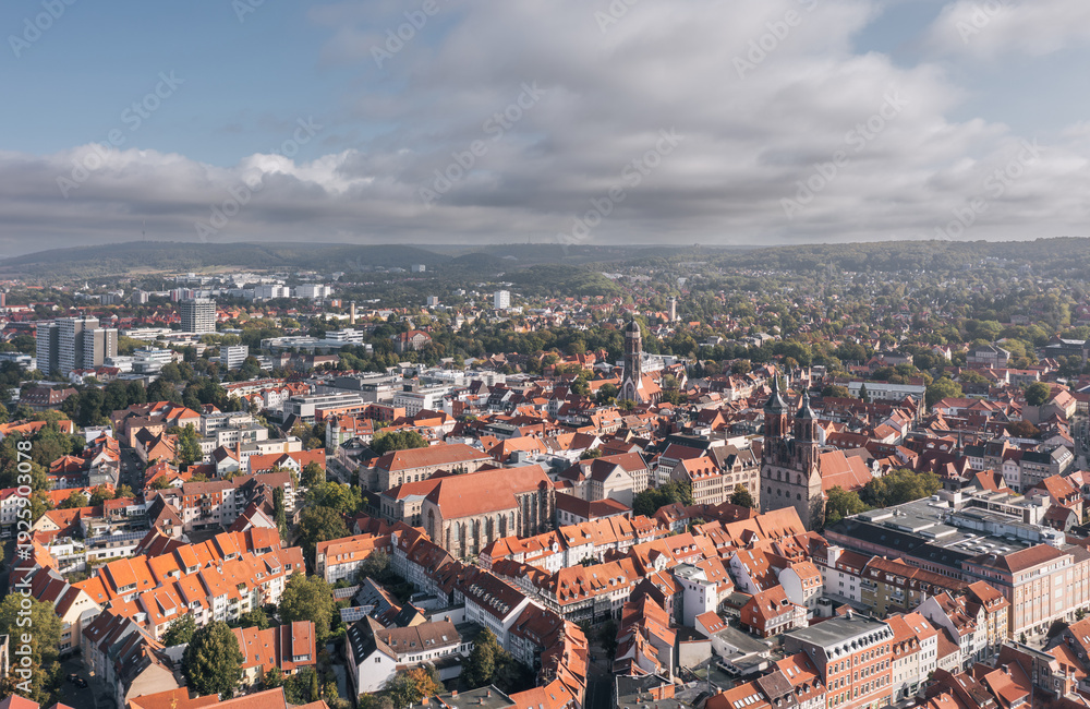 Obraz premium Elevated drone view over the historic old town of Goettingen (Göttingen), Lower Saxony, Germany, featuring dense red rooftops, prominent church towers, and a broad city skyline beneath soft clouds.
