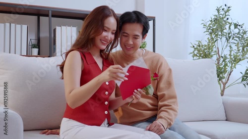 Asian man and girlfriend celebrating Valentine's Day, woman holding card and rose flower with cheerful affection, attractive couple sitting on sofa at home with holiday celebrating and feeling love.