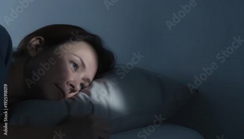 Woman lying awake in bed at night with blue light on face, insomnia and sleep anxiety concept in dark bedroom interior