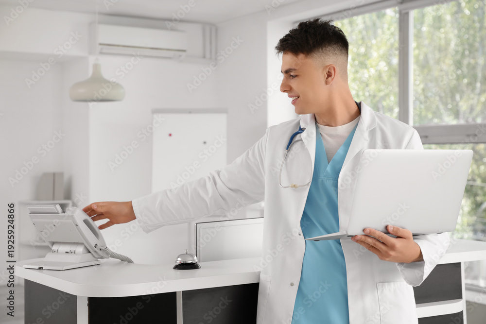 Fototapeta premium Male medical intern with laptop taking phone at reception in clinic