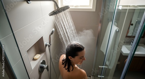 A woman stands under a refreshing shower in a modern bathroom with a glass door and a large rainfall showerhead.