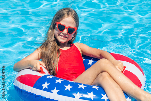 Happy smiling little girl 7-8 years old in red swimsuit and sunglasses relaxing in outdoor swimming pool on lifebuoy decorated with American flag print. Vacation in USA concept. 4th of July pool party