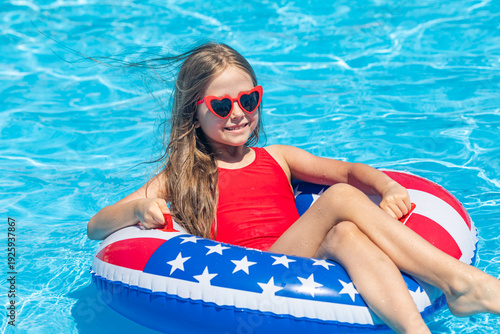 Happy smiling little girl 7-8 years old in red swimsuit and sunglasses relaxing in outdoor swimming pool on lifebuoy decorated with American flag print. Vacation in USA concept. 4th of July pool party