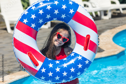 Bild auf Leinwand Happy smiling little girl in red swimsuit and sunglasses stands in outdoor swimming pool, holding a lifebuoy decorated with American flag print