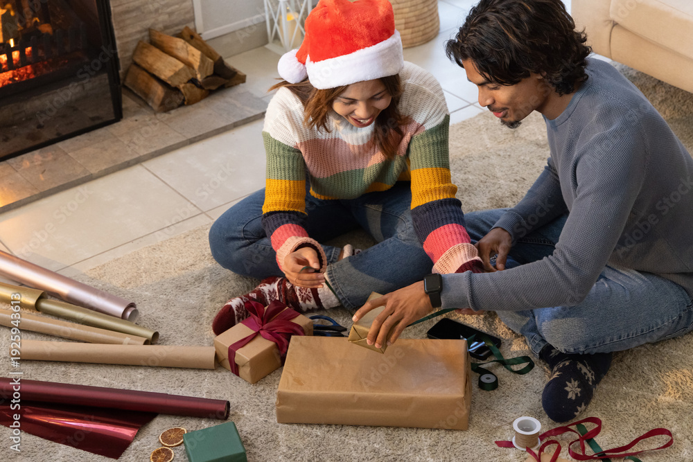 Fototapeta premium Diverse couple sitting on rug by fireplace wrapping gifts with Santa hat and ribbons