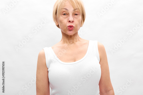 Mature woman in white tank top makes surprised face with puckered lips against plain white background. Concept of emotions, expression, and natural beauty in portrait.