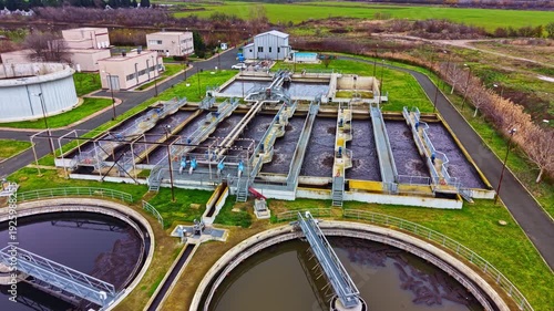 A water treatment facility shows large tanks and processing units. Pipes and machinery are visible. The site is surrounded by open land and trees. The day appears clear.