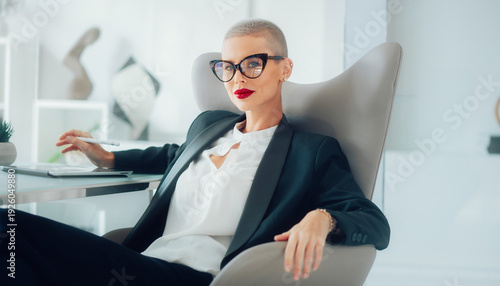 Confident businesswoman with short hair and red lipstick sitting in a modern office chair, holding a pen.