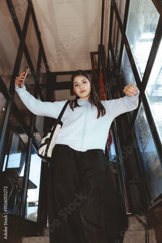 Bild auf Leinwand Confident young Asian woman in casual fashion takes selfie with smartphone while standing on staircase