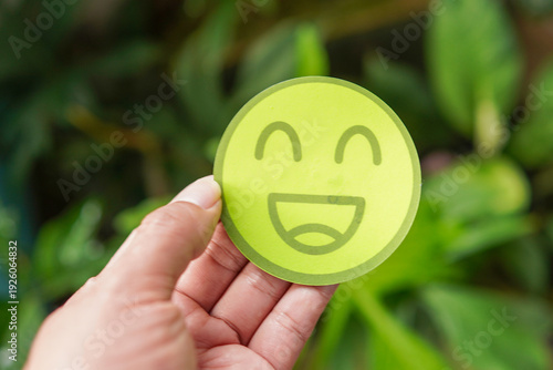 A person's hand holding a bright green paper cut-out with a happy smiling face emoji. The background is a lush, out-of-focus green garden, symbolizing mental health awareness, environmental positivity