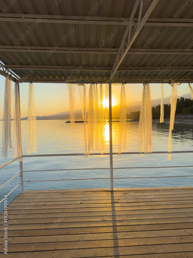 Fototapeta premium Evening sunset at Issyk Kul Lake in Kyrgyzstan. Sun reflecting in calm water viewed from a wooden pier with light curtains.