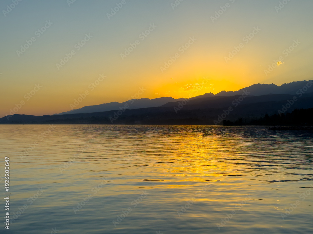 Obraz premium Scenic sunset over Issyk Kul Lake in Kyrgyzstan. The sun sets behind the mountains, casting a golden reflection across the water.