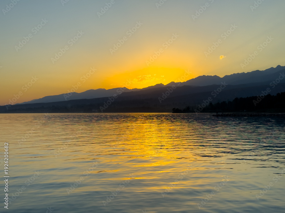 Obraz premium Scenic sunset over Issyk Kul Lake in Kyrgyzstan. The sun sets behind the mountains, casting a golden reflection across the water.