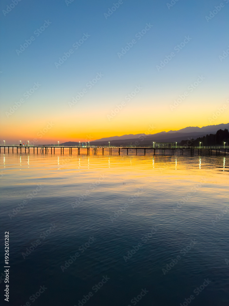 Obraz premium Evening sunset at Issyk Kul Lake in Kyrgyzstan. Long illuminated pier stretching into calm water with mountains on horizon.