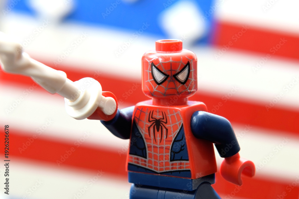 Fototapeta premium Lego minifigure of Spiderman in costume with web in a hand against an American stripped flag. Editorial illustrative image of popular children toys.