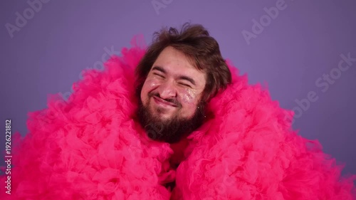 Horizontal 4K video. Young adult man with beard, glitter sparkles on male face, pink tulle fur coat, purple t-shirt looks at camera, smiles, dances, waves his arms, hand pistols on purple background