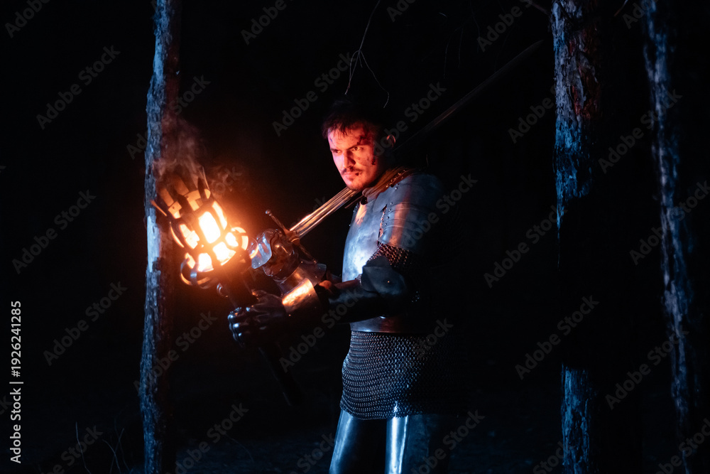 Obraz premium A knight in full armor, sword and torch in hand, walks through a gloomy night forest. A medieval warrior, bloodied and scarred, hunts and searches in the darkness.