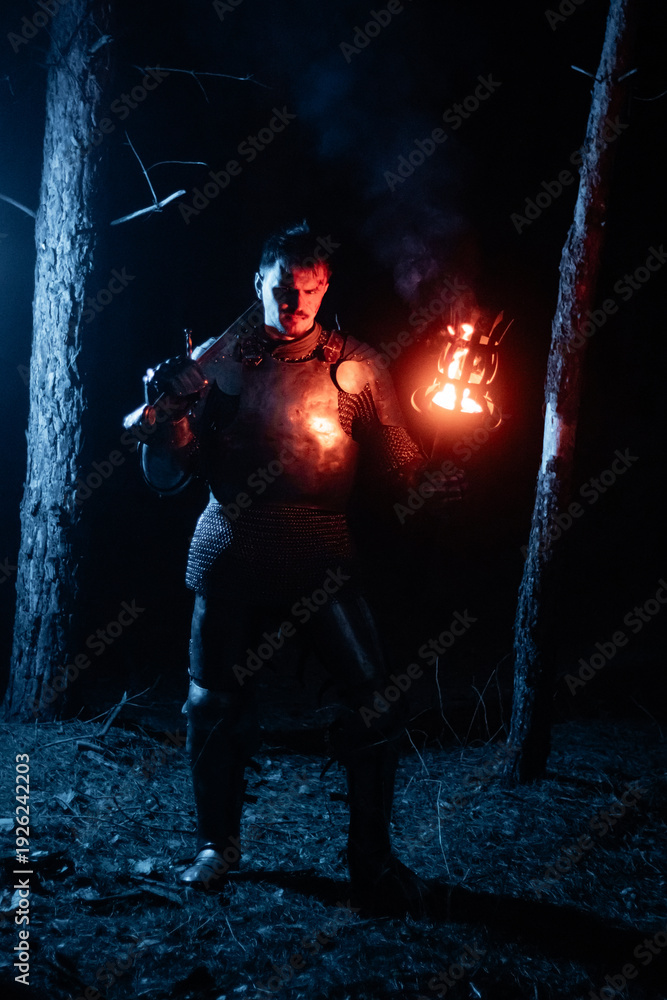 Obraz premium A knight in full armor, sword and torch in hand, walks through a gloomy night forest. A medieval warrior, bloodied and scarred, hunts and searches in the darkness.