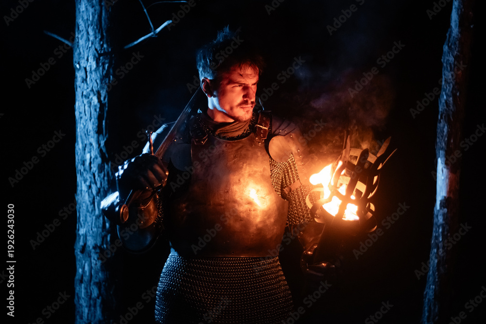 Obraz premium A knight in full armor, sword and torch in hand, walks through a gloomy night forest. A medieval warrior, bloodied and scarred, hunts and searches in the darkness.