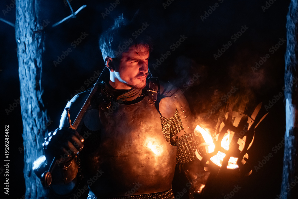 Obraz premium A knight in full armor, sword and torch in hand, walks through a gloomy night forest. A medieval warrior, bloodied and scarred, hunts and searches in the darkness.