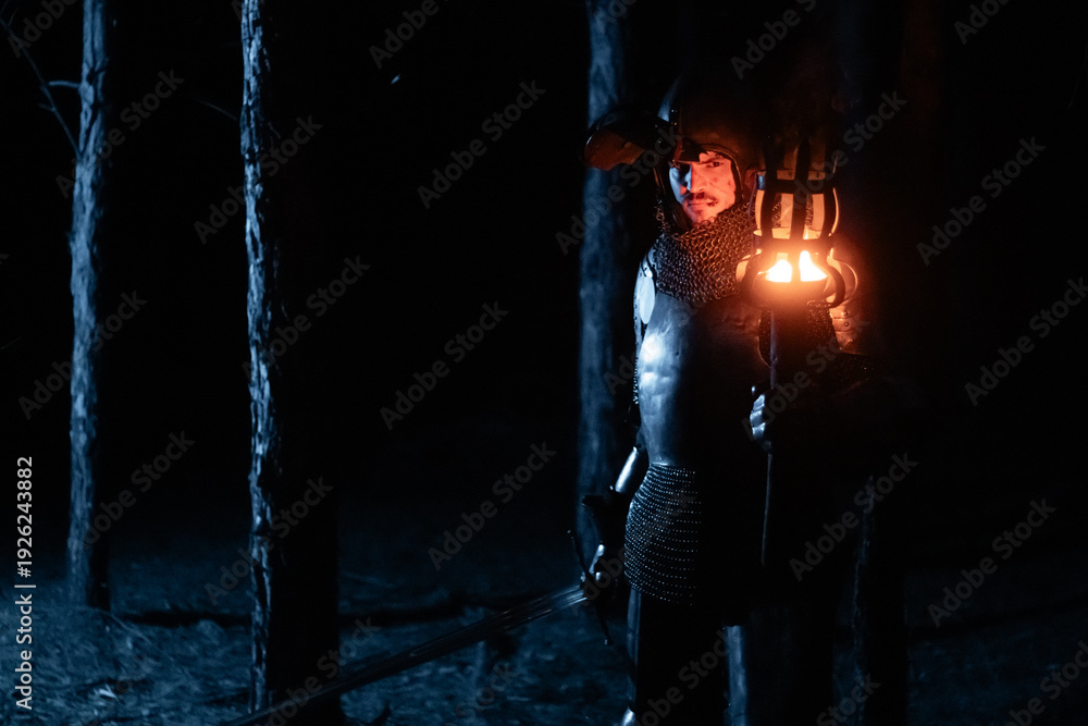 Obraz premium A knight in full armor, sword and torch in hand, walks through a gloomy night forest. A medieval warrior, bloodied and scarred, hunts and searches in the darkness.