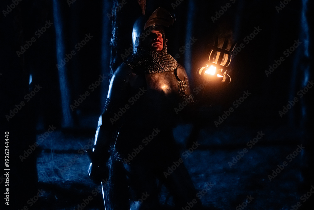 Obraz premium A knight in full armor, sword and torch in hand, walks through a gloomy night forest. A medieval warrior, bloodied and scarred, hunts and searches in the darkness.
