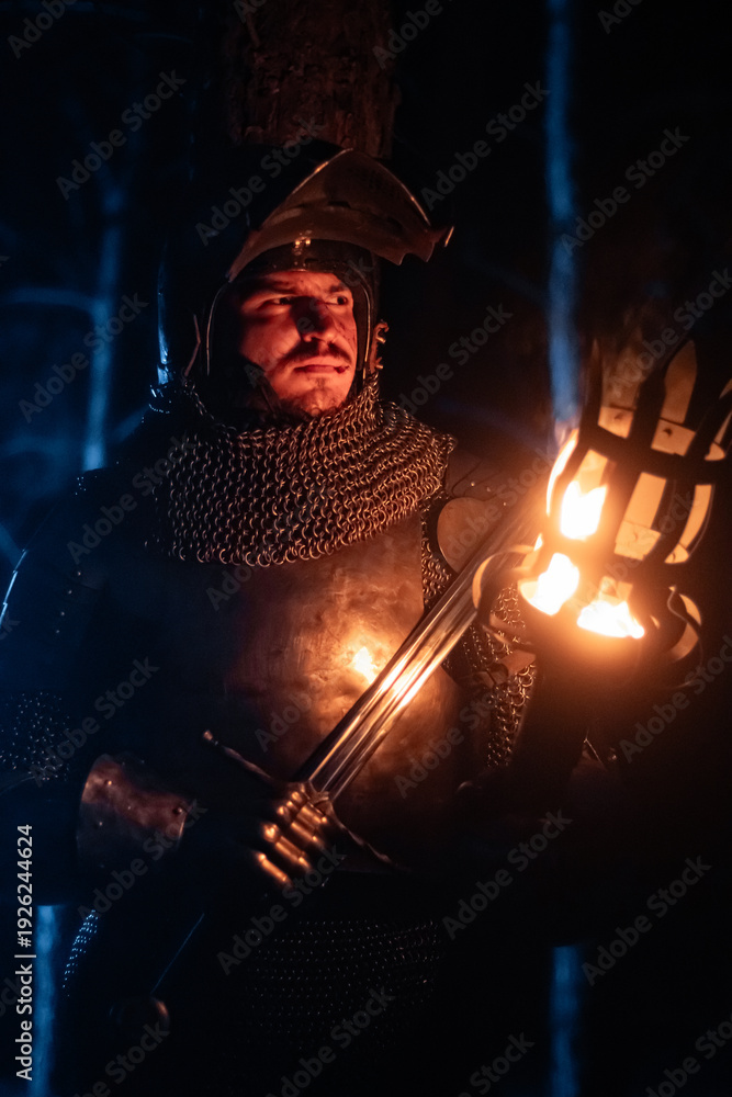 Obraz premium A knight in full armor, sword and torch in hand, walks through a gloomy night forest. A medieval warrior, bloodied and scarred, hunts and searches in the darkness.