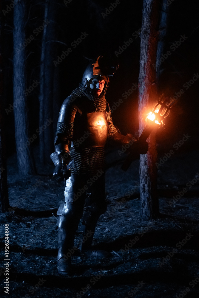 Obraz premium A knight in full armor, sword and torch in hand, walks through a gloomy night forest. A medieval warrior, bloodied and scarred, hunts and searches in the darkness.
