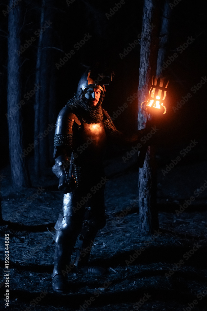 Obraz premium A knight in full armor, sword and torch in hand, walks through a gloomy night forest. A medieval warrior, bloodied and scarred, hunts and searches in the darkness.