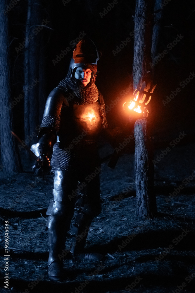 Obraz premium A knight in full armor, sword and torch in hand, walks through a gloomy night forest. A medieval warrior, bloodied and scarred, hunts and searches in the darkness.