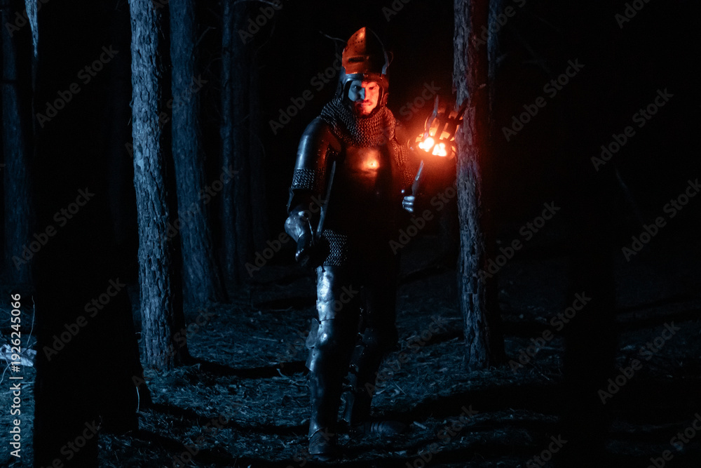Obraz premium A knight in full armor, sword and torch in hand, walks through a gloomy night forest. A medieval warrior, bloodied and scarred, hunts and searches in the darkness.