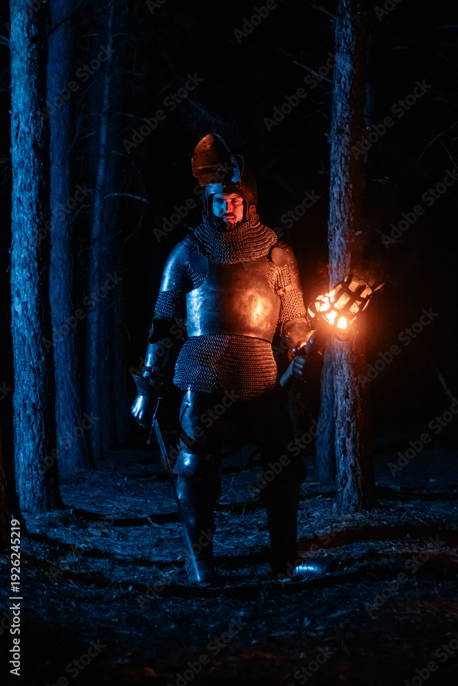 Obraz premium A knight in full armor, sword and torch in hand, walks through a gloomy night forest. A medieval warrior, bloodied and scarred, hunts and searches in the darkness.