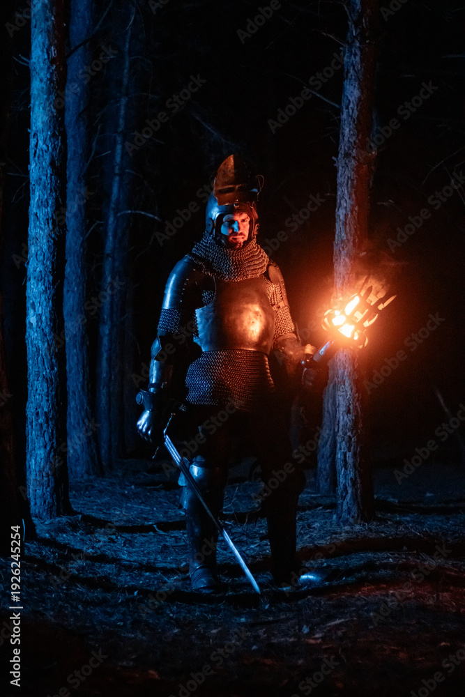Obraz premium A knight in full armor, sword and torch in hand, walks through a gloomy night forest. A medieval warrior, bloodied and scarred, hunts and searches in the darkness.