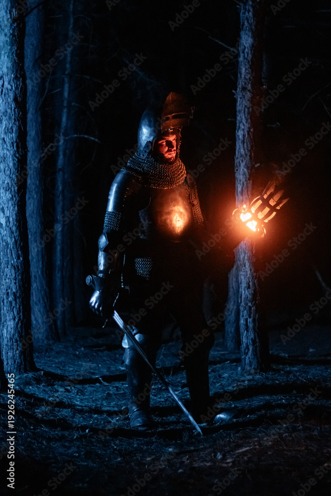 Obraz premium A knight in full armor, sword and torch in hand, walks through a gloomy night forest. A medieval warrior, bloodied and scarred, hunts and searches in the darkness.