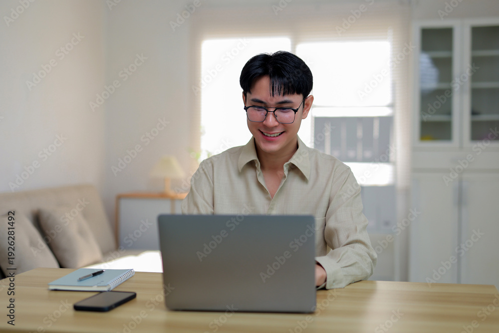 Fototapeta premium Young man wearing glasses smiling while working on a laptop computer at a wooden desk in a modern home office