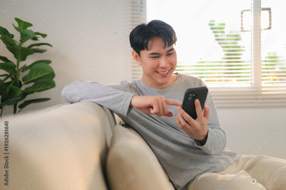 Obraz premium Young man smiling and scrolling on smartphone, connecting with people in a comfortable home environment