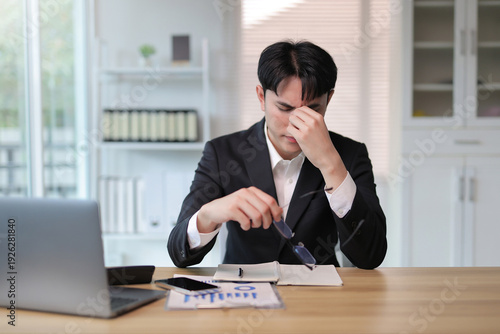 Young businessman experiencing headache and eye strain at office desk, taking off glasses due to overwork and stress