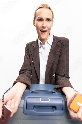 Konstfotografi Surprised woman in brown jacket holding blue suitcase and passport with boarding pass, expressing shock or confusion