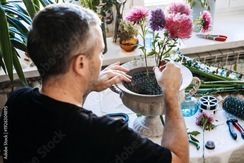 Man inserts pink aster stems into wire mesh inside stone urn on table. male gardener hobby, home grown flowers men, floral lifestyle men.