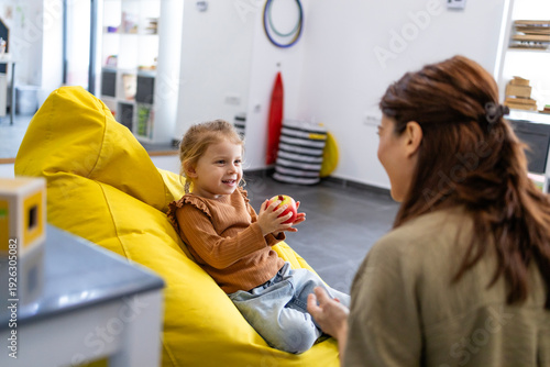 Quadro su tela Young girl on a yellow beanbag holding an apple, smiling and talking with her ch