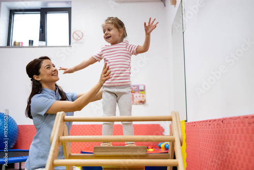 Pediatric physiatrist assisting happy young girl balancing on rehabilitation platform, developing motor skills and coordination during physical therapy session