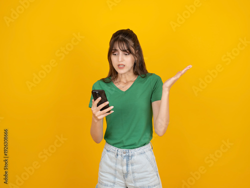 Confused young woman looking at her smartphone with a questioning gesture, standing against a vibrant yellow background. Concept of technological problems, mobile app errors, and feeling lost with dig