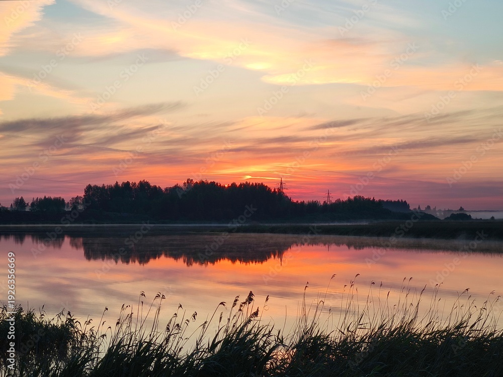 Obraz premium Serene sunset lake wrapped in soft mist rising from the water, glowing with rich orange, pink, violet reflections, surrounded by gentle reeds and dark tree silhouettes under an expressive twiligh sky