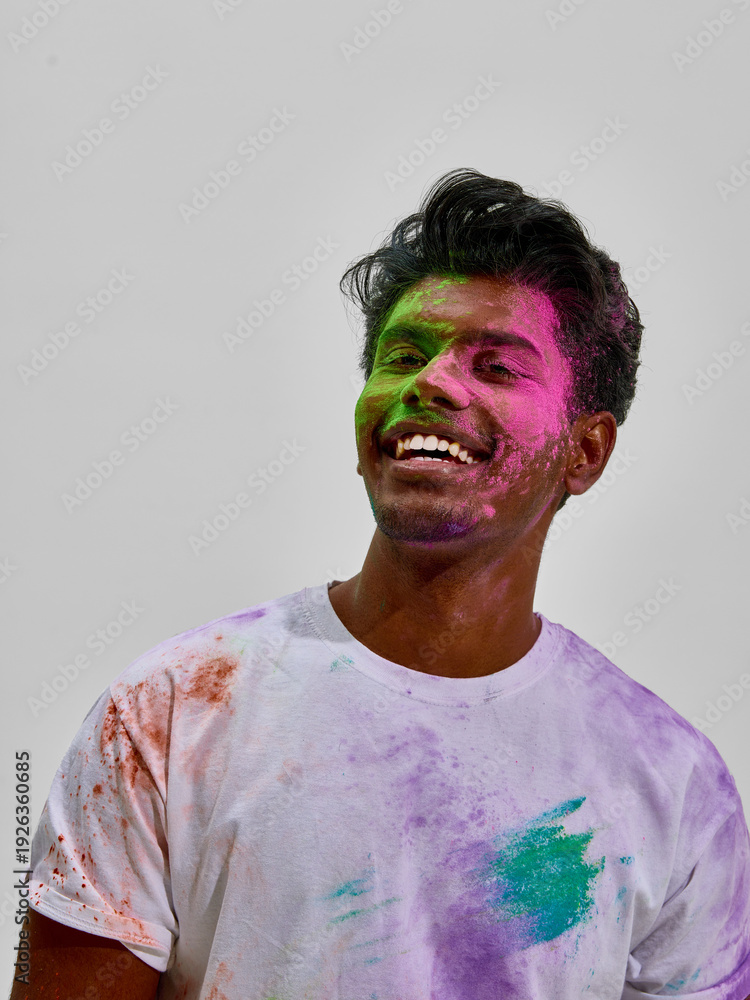Fototapeta premium Young man smiling with colorful powder stains on face and shirt. Concept of festive spring event, casual youth fashion style, cultural celebration atmosphere and lifestyle branding content.