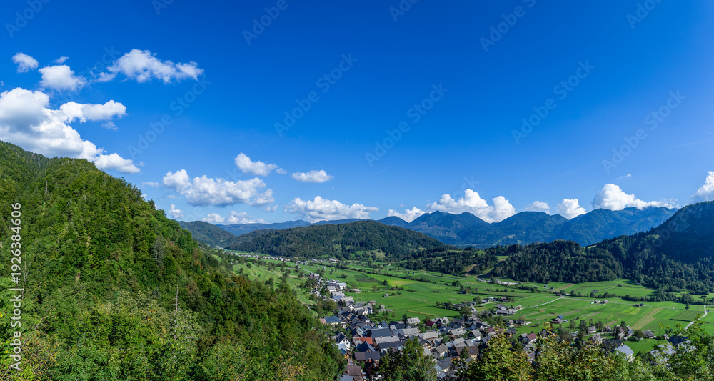 Fototapeta premium Panorama view shows Stara Fuzina in Bohinj valley in Slovenia with green fields and mountains below a blue sky. The area is quiet with no people in sight.