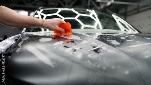 Auto detailer applying paint protection film on a dark car hood with an orange squeegee in a workshop.