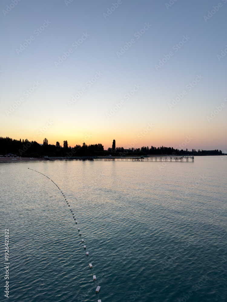 Fototapeta premium Early sunrise at Issyk Kul Lake in Kyrgyzstan. Warm golden sky reflecting in calm water with silhouettes of trees and pier on horizon.